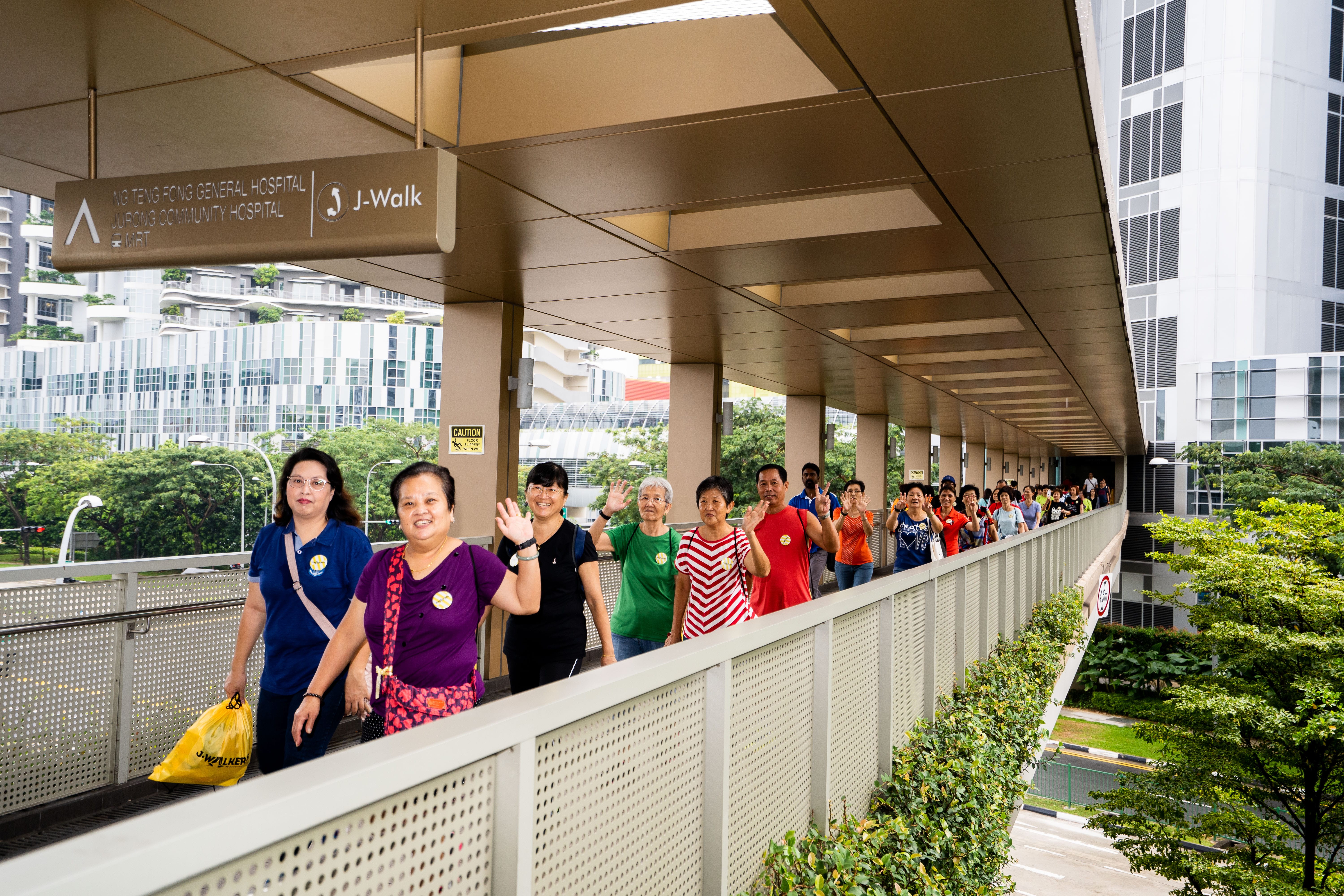 A line of people walking on a covered pedestrian bridge with greenery and buildings visible in the background.